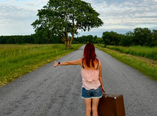 A woman with red hair hitchhiking on a country road in Estonia with a vintage suitcase.