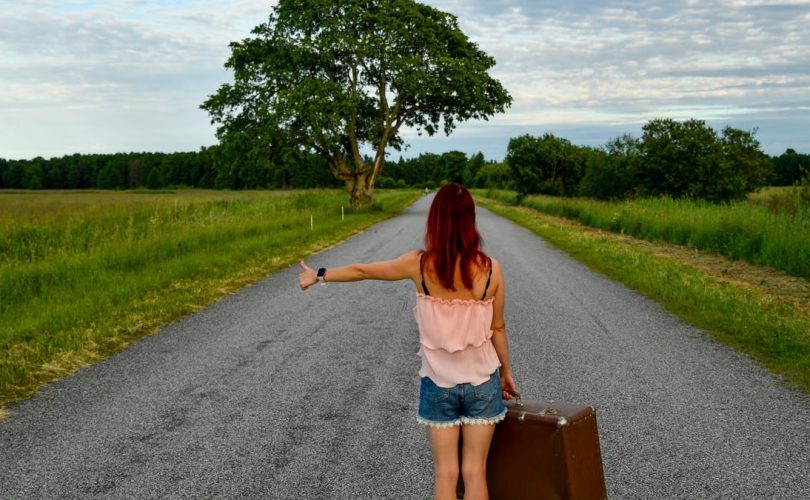 A woman with red hair hitchhiking on a country road in Estonia with a vintage suitcase.